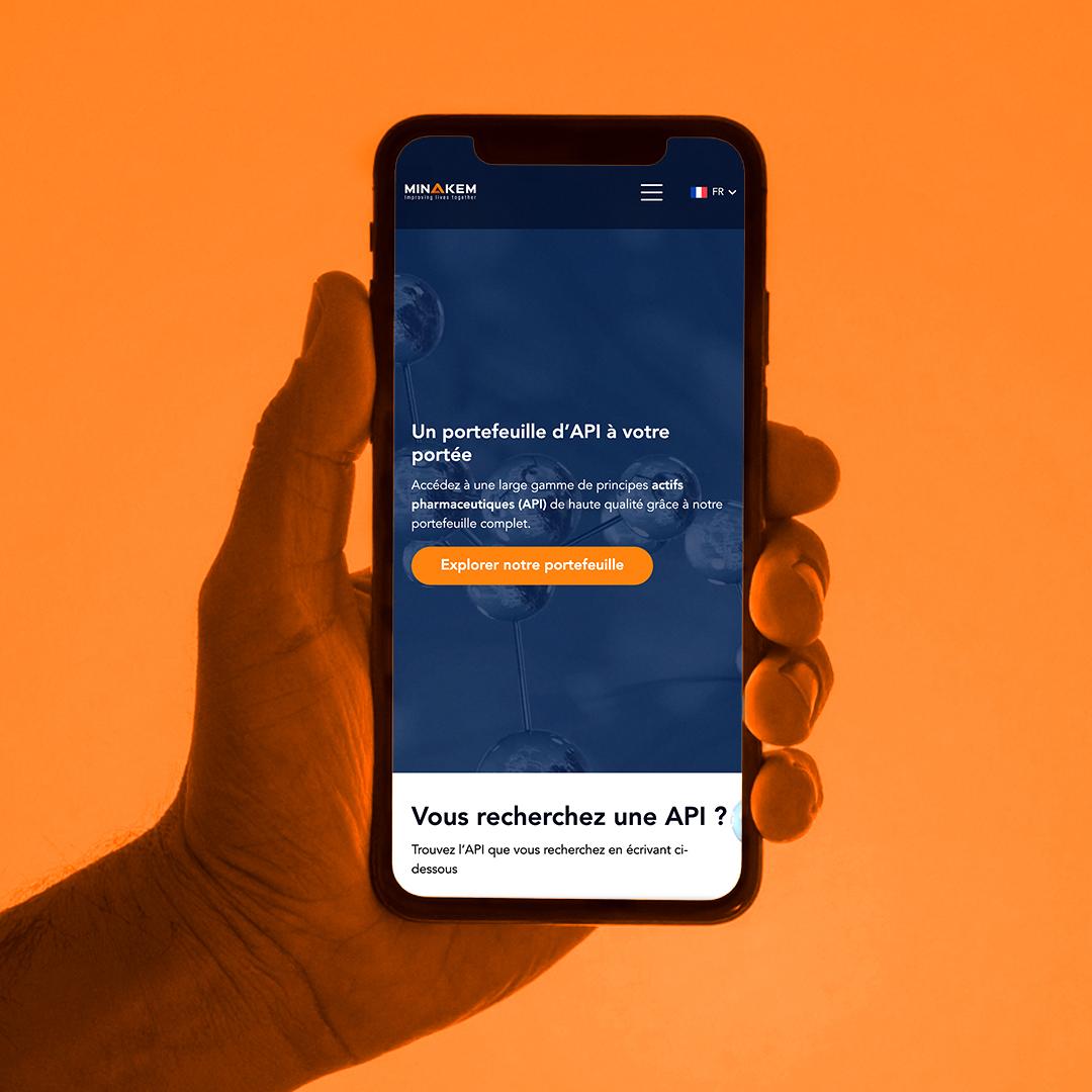 Smartphone tenu en main affichant la page mobile Minakem avec un bouton “Explorer notre portefeuille”